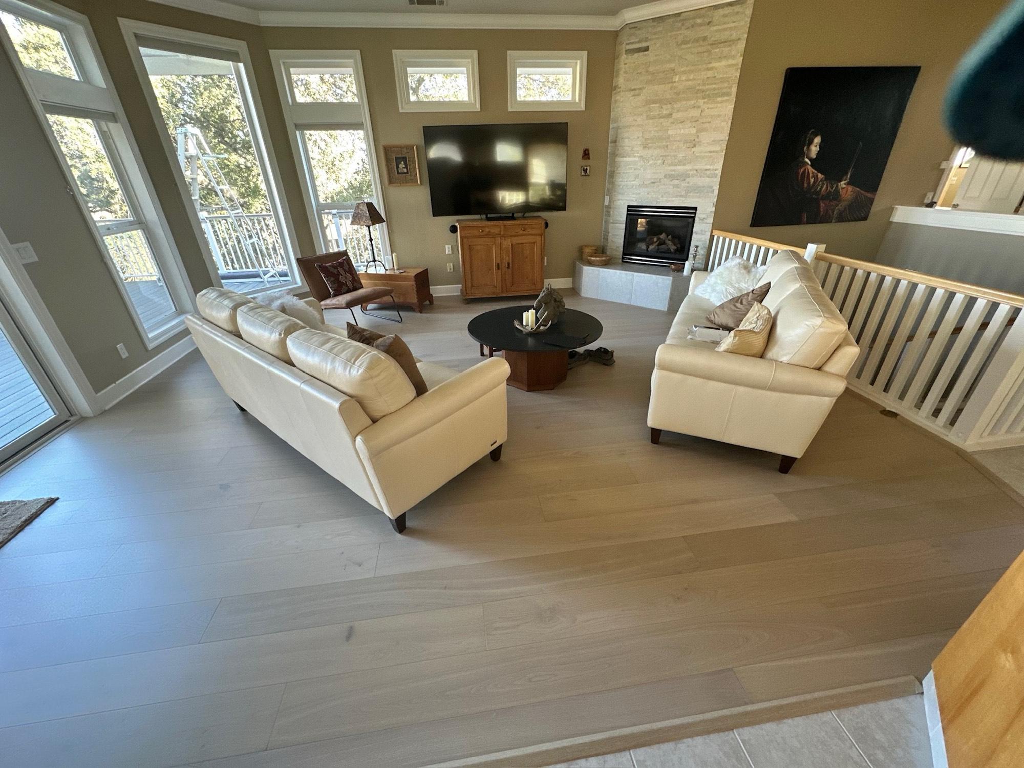 Wide-plank hardwood floor in a spacious living room with a stone fireplace and floor-to-ceiling windows.