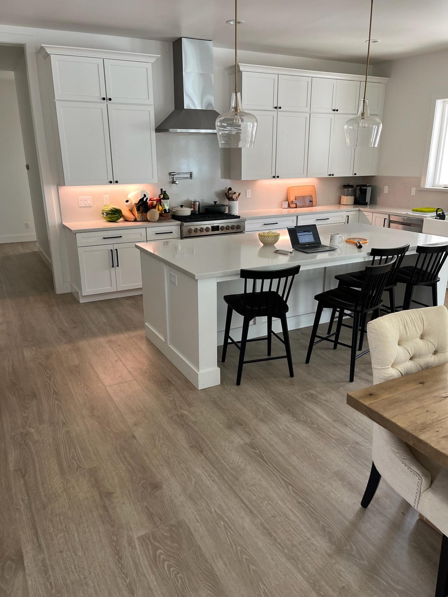 Modern white kitchen with a large island, pendant lights, and light gray wide-plank vinyl flooring.